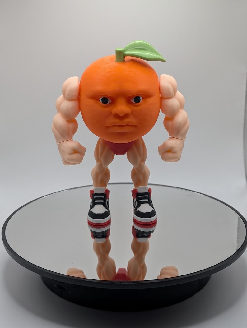 Puede incluir: Figura de naranja impresa en 3D con rostro humano, brazos y piernas musculosos. La naranja tiene una hoja verde y lleva calzoncillos rojos y zapatillas deportivas blancas y negras. La figura est&aacute; sobre una superficie reflectante.