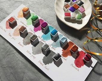Pode incluir: Um conjunto de 12 tintas de aquarela em várias cores, incluindo manga, verde, magenta, verde azulado, azul seda, tingido, fúcsia, vinho tinto, ouro rosa, marrom, cinza prateado e pérola preta. As tintas estão dispostas em uma fileira sobre uma superfície branca.