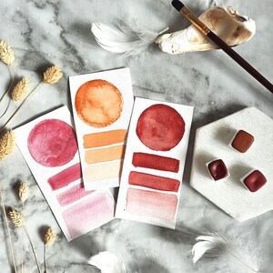 May include: Three watercolour paint swatches on white paper with a red, orange, and pink colour scheme. The swatches are arranged in a vertical column. The paper is on a marble surface.