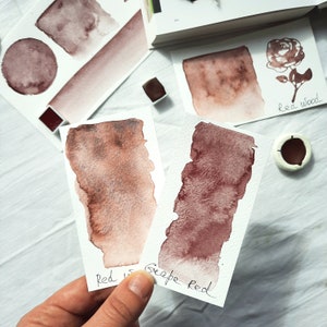 May include: Two watercolor paint swatches on white paper. The swatch on the left is labeled "Red", and the swatch on the right is labeled "Cape Red".