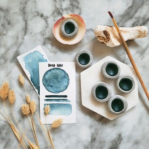 Può includere: Un set di colori ad acquerello con un campione di lavaggio ad acquerello blu e il testo "Deep Lake". Il set comprende sei piccoli piatti di pittura in ceramica rotondi con una smaltatura blu scuro.
