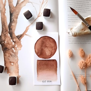 Op de afbeelding: Een aquarelverfpan met het label "VELVET BROWN", een bruin verfstaal en een cirkel van bruine verf. De verfpan ligt op een wit oppervlak met een boek en gedroogde bloemen.