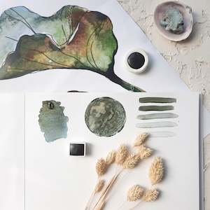 May include: A watercolor painting of a leaf with a green, brown, and gray color scheme. The painting is on a white background. There are also swatches of green and gray watercolor paint on a white sheet of paper. A small, square, black paint pan is also visible.