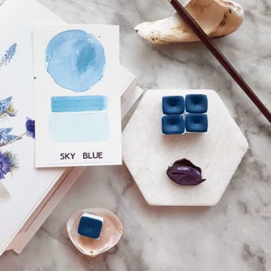May include: A watercolour paint sample card with the text "SKY BLUE" and a blue watercolour wash. Four blue watercolour paint pans are arranged in a square on a white hexagonal tile. A single blue watercolour paint pan is in a white seashell.