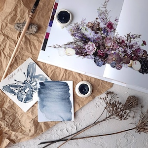 May include: A watercolour painting of a butterfly with the text "Night Sky" on a white card. There are two small ceramic dishes with black paint in them. The dishes are on a brown paper surface.