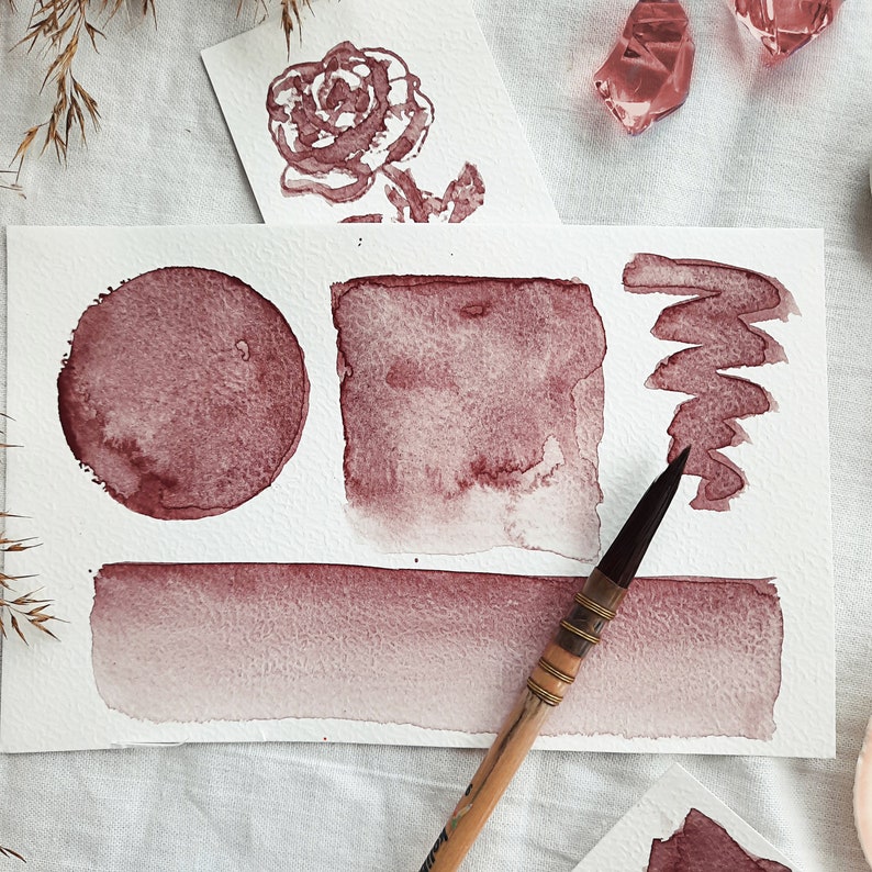 May include: A watercolor painting on white paper with a brush. The painting features a circle, a square, a rectangle, and a zig-zag line. The shapes are painted in shades of red.
