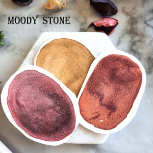 Può includere: Tre campioni di acquerello su una superficie bianca. I campioni sono in tonalità di marrone, rosso e arancione. Il testo "MOODY STONE" è scritto sopra i campioni.