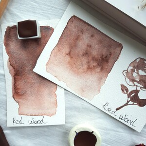 May include: Two watercolor swatches of a reddish-brown color labeled "Red Wood". The swatches are on white paper and the paint is in a pan.