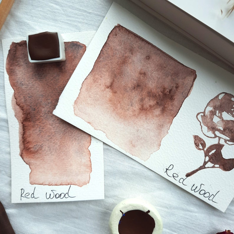 May include: Two watercolor swatches of a reddish-brown color labeled "Red Wood". The swatches are on white paper and the paint is in a pan.