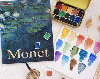 May include: A book titled "Monet" with a painting of water lilies on the cover. A small tin of watercolor paints with a variety of colors is open next to the book. A sheet of paper with watercolor swatches is on the table. A paintbrush and a few other watercolor supplies are also on the table.