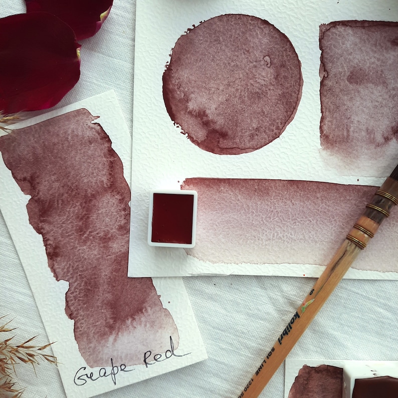 May include: A watercolor paint pan with a red paint sample, labeled "Grape Red". The image also shows several watercolor washes in different shades of red on white paper.