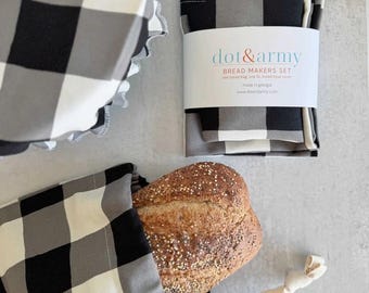 Puede incluir: Un juego de panadería con un patrón de cuadros negros y blancos. El set incluye una funda de tela para cuencos con borde fruncido y una bolsa para pan con cordón que contiene una hogaza de pan con semillas. Una etiqueta blanca dice dot & army.