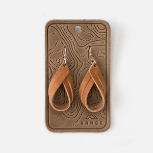 May include: A pair of brown leather earrings with a twisted design, hanging from silver hooks. The earrings are displayed on a brown leather card with a topographic map pattern and the word "RANGE" printed on the bottom.