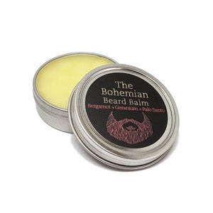 May include: A silver tin with a yellow beard balm inside. The label on the tin reads "The Bohemian Beard Balm Bergamot + Geranium + Palo Santo" and features a black and red illustration of a beard.