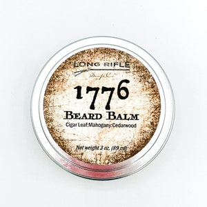 May include: A silver tin with a label that reads "LONG RIFLE 1776 BEARD BALM Cigar Leaf:Mahogany:Cedarwood Net weight 3 oz. (89 ml)"