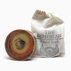 May include: A round, multi-colored beard soap bar with a swirl pattern in shades of brown, orange, and yellow. The soap is packaged in a white muslin drawstring bag with the text "THE BOHEMIAN  BERGAMOT + GERANIUM + PAO SANTO BEARD SOAP" printed on the front.
