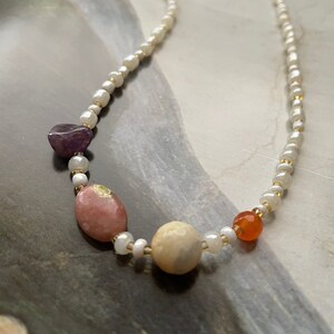 May include: A white pearl necklace with a gold clasp and four gemstone beads: a purple amethyst, a pink and white stone, a cream-colored stone, and an orange carnelian.