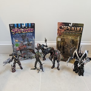 May include: A collection of Spawn action figures, some still in their original packaging. The figures are of various characters, with detailed designs and weaponry. The packaging includes the text "Spawn Ultra-Action Figures" and "Spawn The Dark Ages".