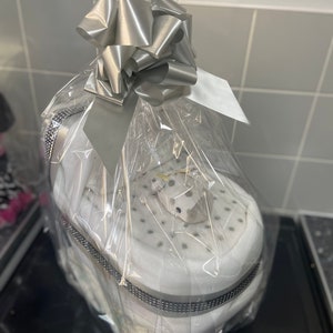 May include: A white baby shoe gift basket wrapped in clear cellophane with a silver bow. The shoe is decorated with silver glitter and rhinestones.