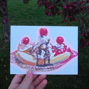 May include: A vibrant illustration of a banana split against a light blue backdrop. The dessert features bananas, ice cream, whipped cream, chocolate sauce, and cherries. The artwork is held by a hand, set against green grass and red-flowered trees.