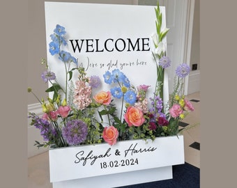 May include: A white welcome sign with a floral arrangement in front of it. The sign says "Welcome" and "We're so glad you're here". Below the welcome message is a white box with the names "Safiyah & Harris" and the date "18.02.2024".