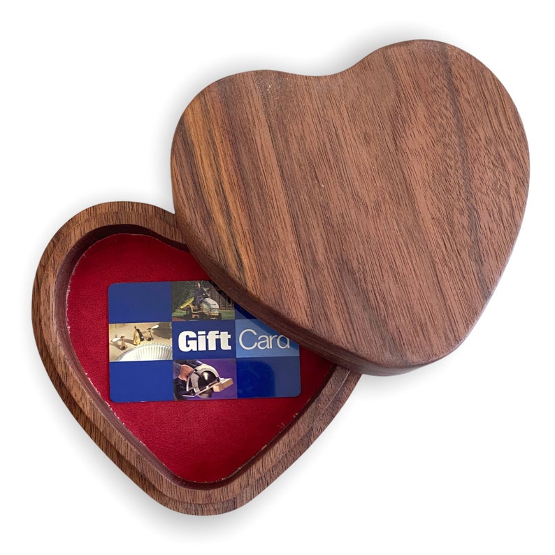 Puede incluir: Una caja de madera en forma de coraz&oacute;n con un acabado de veta de madera marr&oacute;n oscuro y una tapa parcialmente abierta. El interior de la caja est&aacute; forrado con tela roja y contiene una tarjeta de regalo rectangular con paneles azules.