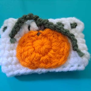 May include: White crocheted cup cozy with an orange crocheted pumpkin design and green crocheted stem.