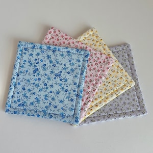 May include: Five fabric coasters with floral patterns. The coasters are layered on top of each other, with a blue floral coaster on the bottom, a pink floral coaster in the middle, and a yellow floral coaster on top. The coasters are all made of a soft, absorbent fabric.