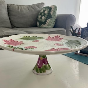 May include: White ceramic cake stand with a scalloped edge, featuring pink and green floral designs. The pedestal base is painted with pink and green accents. A decorative piece for displaying baked goods or other items.