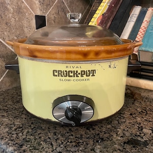 May include: A pale yellow Rival Crock-Pot slow cooker with a brown ceramic rim and a clear glass lid. The front features a black control knob with silver accents labeled OFF, LOW, and HIGH. The brand name is printed in black on the front surface.