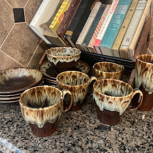May include: A collection of brown and cream-colored ceramic tableware, including plates, bowls, and mugs. The mugs have handles and a unique glaze pattern. A tall tumbler is also included. The set is arranged on a speckled countertop.