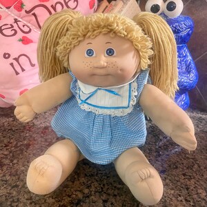 May include: A doll with blonde yarn hair styled in two pigtails, blue eyes, and freckles. The doll wears a light blue and white gingham dress with a white collar featuring blue trim and a small blue ribbon bow at the center of the neckline.