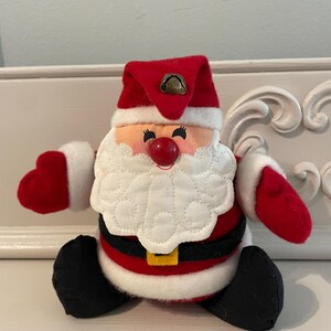 May include: A plush Santa Claus figure featuring a red hat with a small metal bell, a white quilted beard, a round red nose, and black eyes. The figure wears a red suit with white trim, a black belt with a yellow buckle, and black boots.