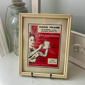 May include: A rectangular off-white picture frame containing a vintage advertisement insert. The insert features red and black text, including E-Z HANG FRAME, and a black and white illustration of a person holding a frame. It states Size: 7 x 9.