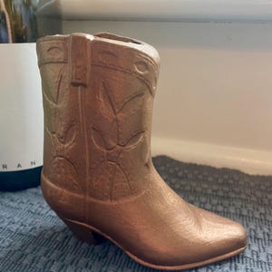 May include: A bronze-colored cowboy boot-shaped vase. The boot features embossed floral designs and a pointed toe. The vase has a small heel and is made of a solid material.