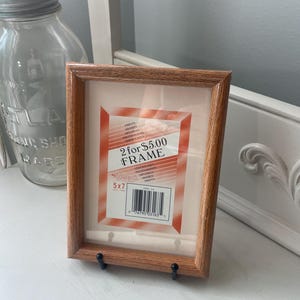 May include: A rectangular wooden picture frame with a light brown grain finish. The frame contains a white insert with orange corner accents and text that reads 2 for $5.00 FRAME, 5x7, and Intercraft. The frame is supported by two small black stands.