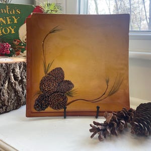 May include: A square, vintage-style painting with a warm, golden-brown background. The artwork features a cluster of detailed pine cones and delicate green pine needles. The painting is propped up on a black stand, with additional pine cones nearby.
