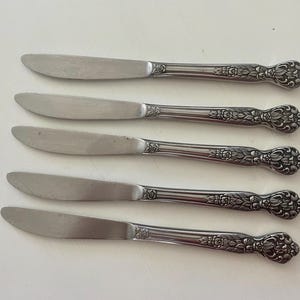 May include: Five stainless steel butter knives with ornate floral detailing on the handles. The blades are smooth and curved, and the handles have a decorative, vintage design. The knives are arranged on a white surface.