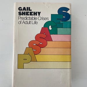 May include: A vintage book titled "Predictable Crises of Adult Life" by Gail Sheehy. The cover features the word "PASSAGES" in colorful, stacked letters over a series of horizontal stripes in shades of yellow, orange, pink, green, and blue.