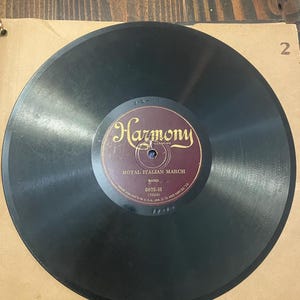 May include: A vintage black vinyl record with a maroon and gold label. The label reads "Harmony" and "Royal Italian March." The record is in a cardboard sleeve. The record's diameter is approximately 12 inches.