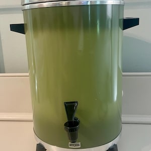 May include: A cylindrical olive green West Bend percolator with a silver metal lid and base. It features black plastic side handles, a black plastic spigot on the front, and three black plastic legs. The West Bend logo is printed on a small silver plate.