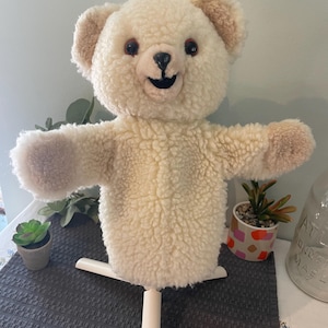May include: A plush, cream-colored teddy bear puppet with a smiling face, brown eyes, and tan accents on the ears and paws. The puppet is mounted on a white stand, with its arms outstretched. The bear is made of a soft, textured material.