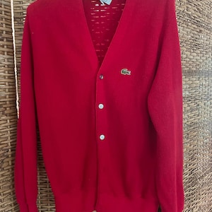 May include: A bright red cardigan sweater with a V-neck and button closure. The sweater features a small green crocodile logo on the chest. The collar is decorated with a rainbow-colored trim. The sweater is made of a soft material.