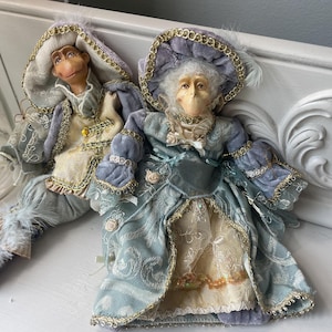 May include: Two collectible dolls dressed in elaborate period-style clothing. The left doll wears a light blue and cream velvet outfit with gold trim. The right doll wears a blue velvet gown with a matching bonnet, lace details, and gold accents.