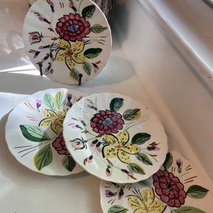 May include: A set of four decorative ceramic plates with floral designs. Each plate features a central arrangement of yellow and maroon flowers with green leaves against a white background. The plates are displayed on a white surface, with one plate on a black stand.
