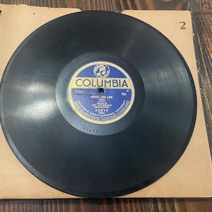 May include: A vintage black vinyl record with a blue and yellow label, featuring the text "Columbia" and "Sweet and Low." The record is housed in a tan paper sleeve, with the number "2" visible. The record is from Art Hickman's Orchestra.