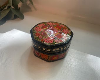May include: A small, black, octagonal box with a floral design on the lid. The lid features a pink and red floral pattern with green leaves. The box has gold accents and sits on a white surface. Decorative storage.