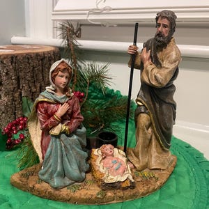 May include: A detailed nativity scene figurine set. The figures include Mary, Joseph, and baby Jesus in a manger. The figures are painted in muted tones with gold accents. The scene is set on a base with greenery and a small black bucket.