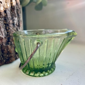 May include: A small, light green glass bucket with a silver metal handle. The bucket has a ribbed design and a decorative base. The bucket is likely vintage or antique and is displayed on a white surface.