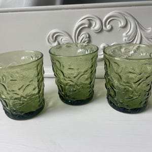 May include: Three olive green textured glass tumblers. The glasses have a unique, organic pattern and a dark green base. The tumblers are arranged on a white surface, with a decorative white background.